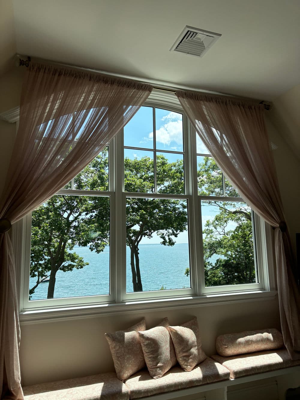 Lakeview window with sheer curtains and decorative cushions, framed by lush trees.